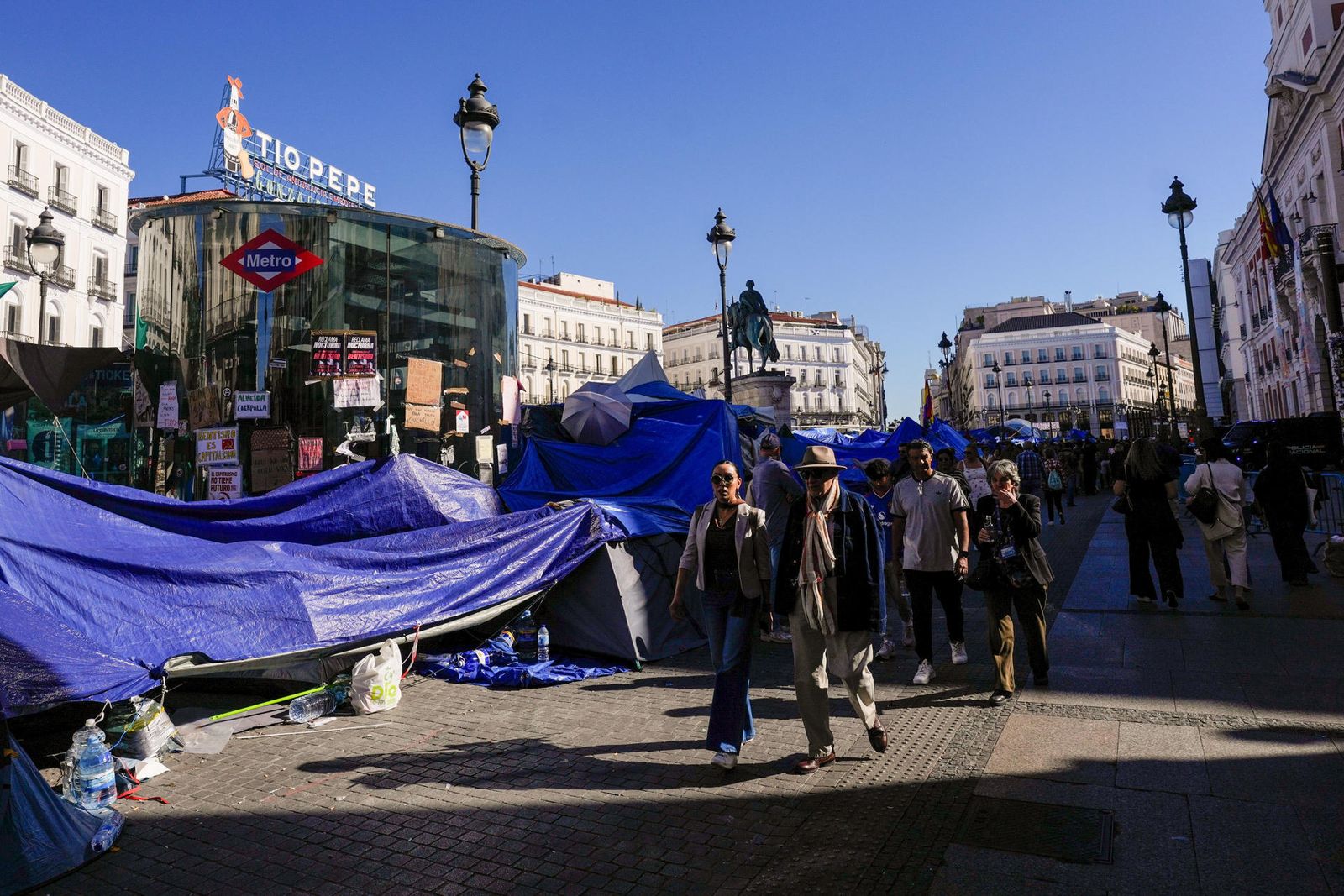 Transeúntes caminan junto a la acampada protesta contra la crisis de la vivienda en la Puerta del Sol