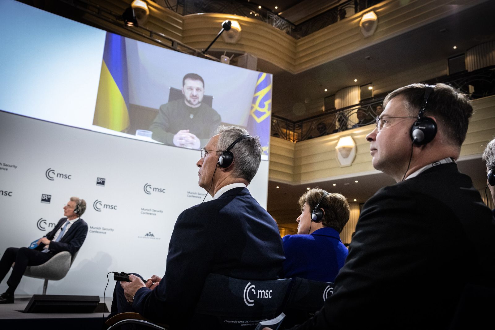 El Secretario General de ATO, Jens Stoltenberg (C), Christoph Heusgen (L), Presidente de la Conferencia de Seguridad de Munich, y el Comisario Europeo de Comercio, Valdis Dombrovskis (R), escuchan al presidente ucraniano Volodymyr Zelensky (pantalla pn) durante la 59.ª Conferencia de Seguridad de Munich