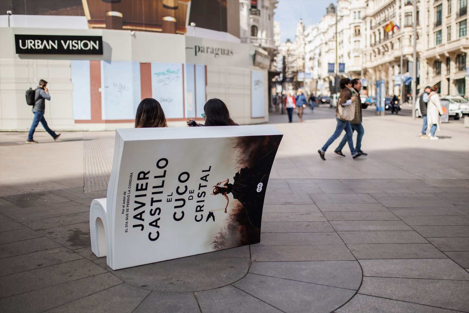 Dos personas se sientan en un banco con forma de libro de la campaña ‘Asienta la lectura-siéntate a leer’, en Gran Vía