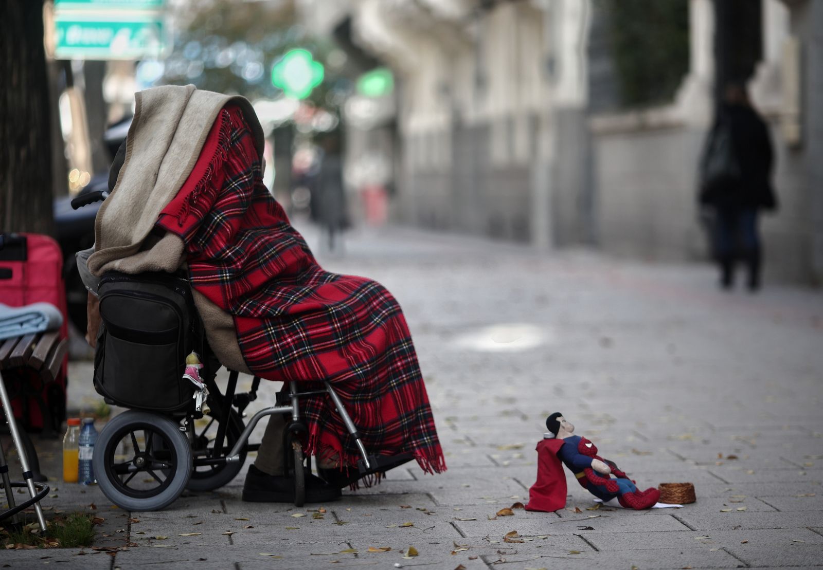 Una persona en silla de ruedas y tapada con una manta pide dinero con un muñeco de Superman en una céntrica calle