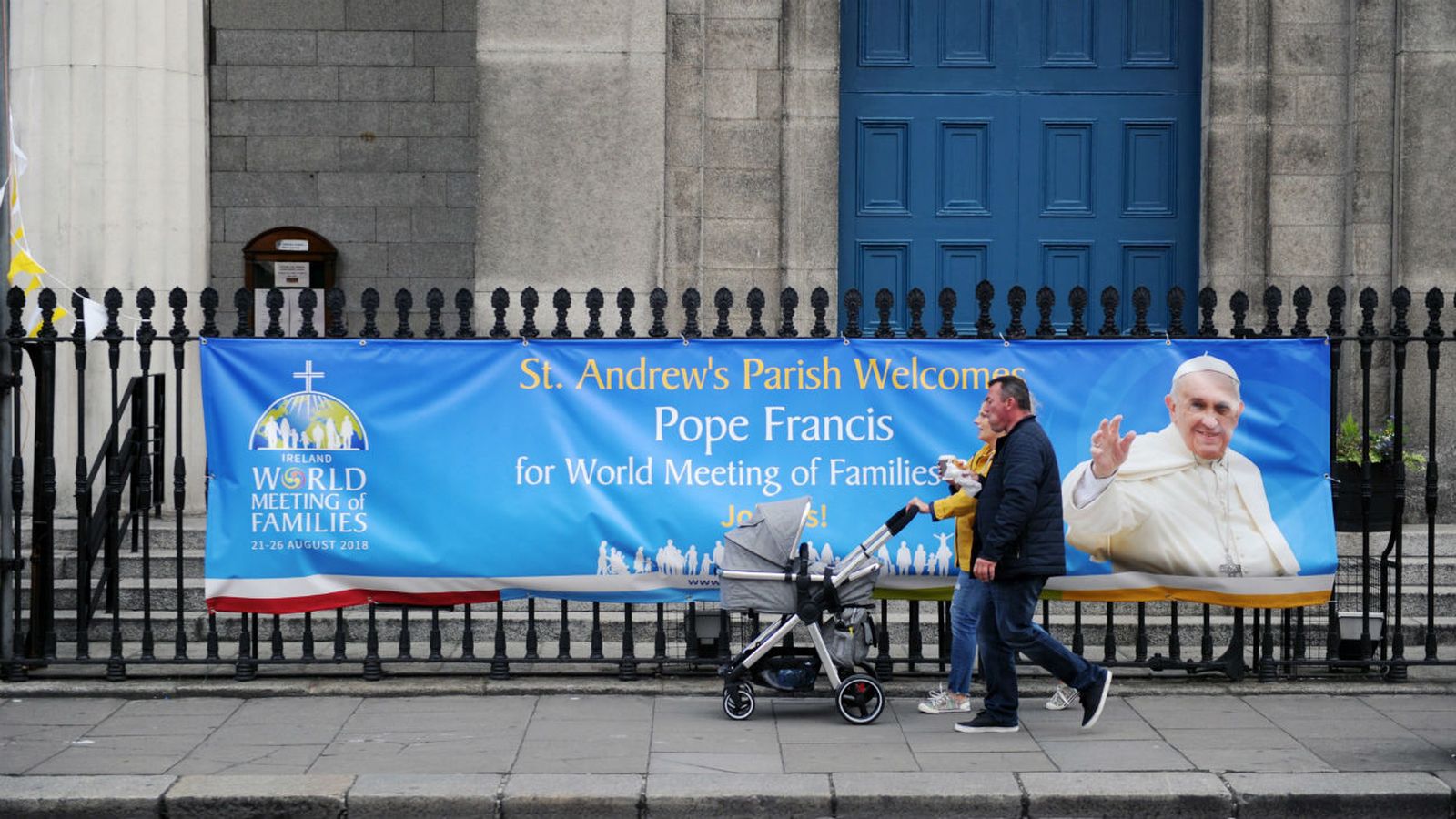 Una familia camina delante de una pancarta con la imagen del papa Francisco situada fuera de una iglesia en Dublín