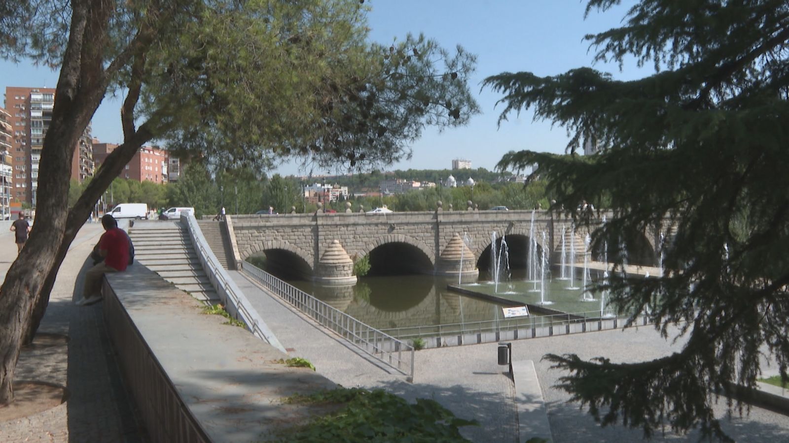 Dos personas se protegen del calor bajo la sombra de un árbol en el entorno de Madrid Río